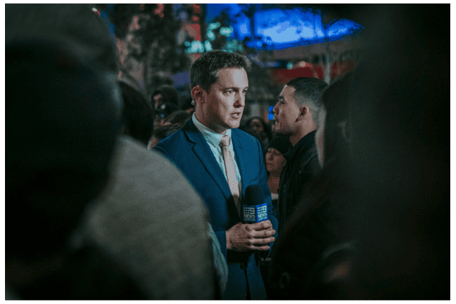 A man in a blue suit holds a microphone and speaks in front of a crowd.